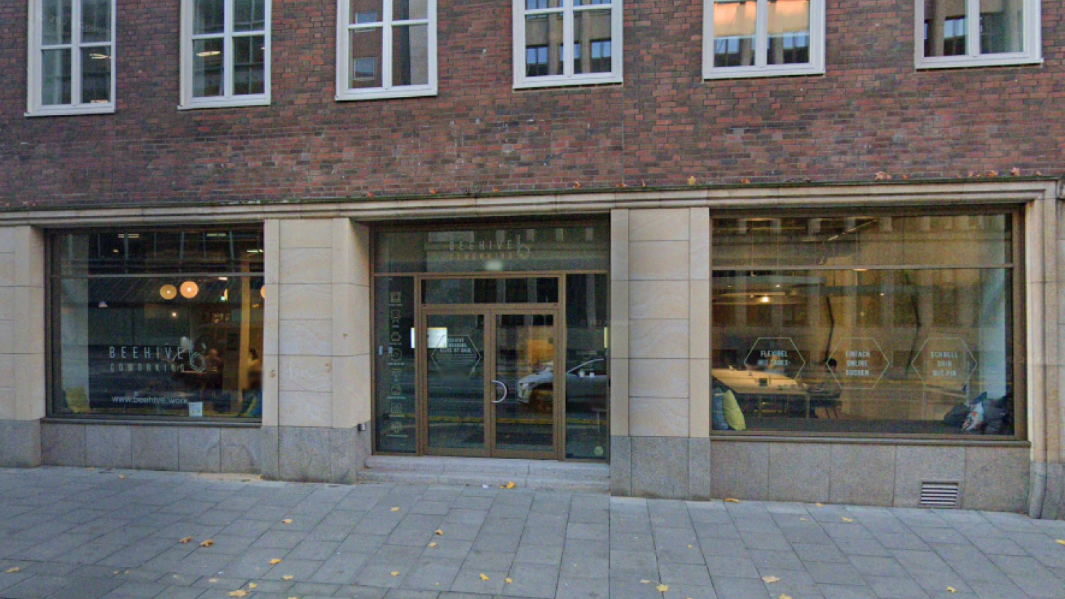  Straßenansicht eines Coworking-Spaces mit großer Glasfassade in einem Backsteingebäude in Hamburg; auf den Fenstern sind Begriffe wie „flexibel“ und „einfach online buchen“ zu lesen.