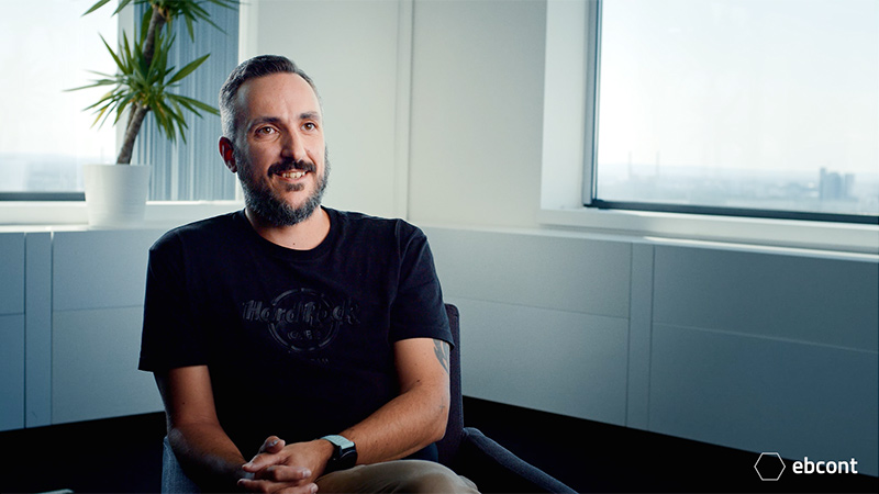 Ausschnitt aus dem Video auf dem Mitarbeiter Tom entspannt in einem hellen Raum sitzt und lächelt in die Kamera. Er trägt ein schwarzes T-Shirt.