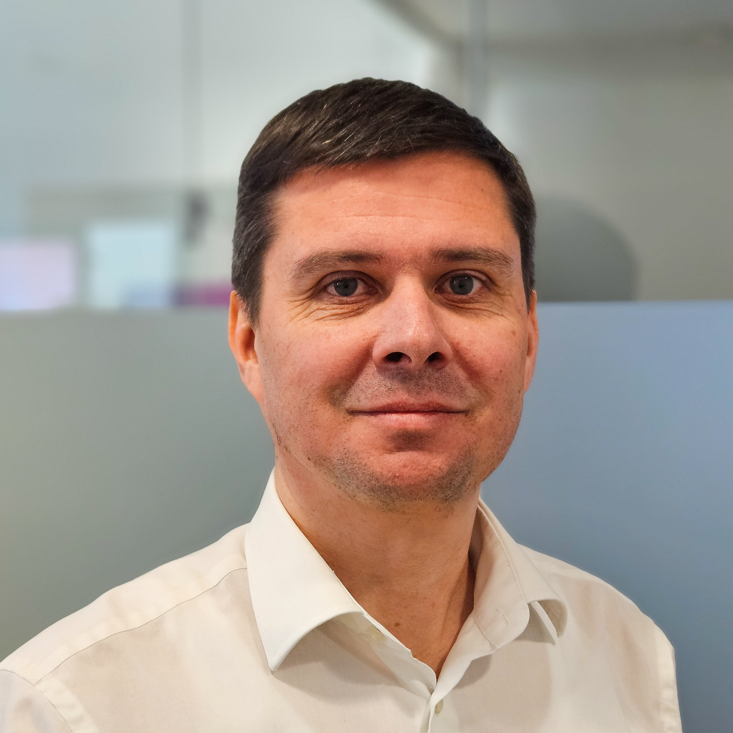 Mann mit kurzem, dunklem Haar trägt ein weißes Hemd und blickt freundlich in die Kamera. Der Hintergrund ist ein modernes Büro mit Glaswänden und matter Abtrennung.