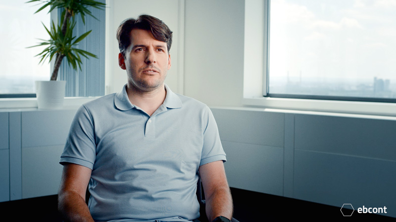 Ausschnitt aus dem Video auf dem Mitarbeiter József entspannt in einem hellen Raum sitz. Er trägt ein hellgraues Polo T-Shirt.