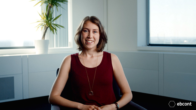 Ausschnitt aus dem Video auf dem Mitarbeiterin Irene entspannt in einem hellen Raum sitzt und ind die Kamera lächelt. Sie trägt ein dunkelrotes Top.