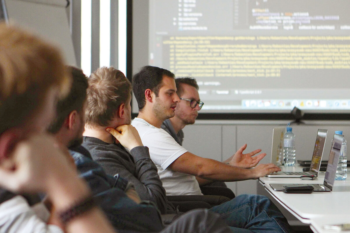 Several young men are sitting at a long table with laptops in a modern meeting room. One of them, wearing a white T-shirt, is gesticulating and talking. A large screen with code and a development environment can be seen in the background.