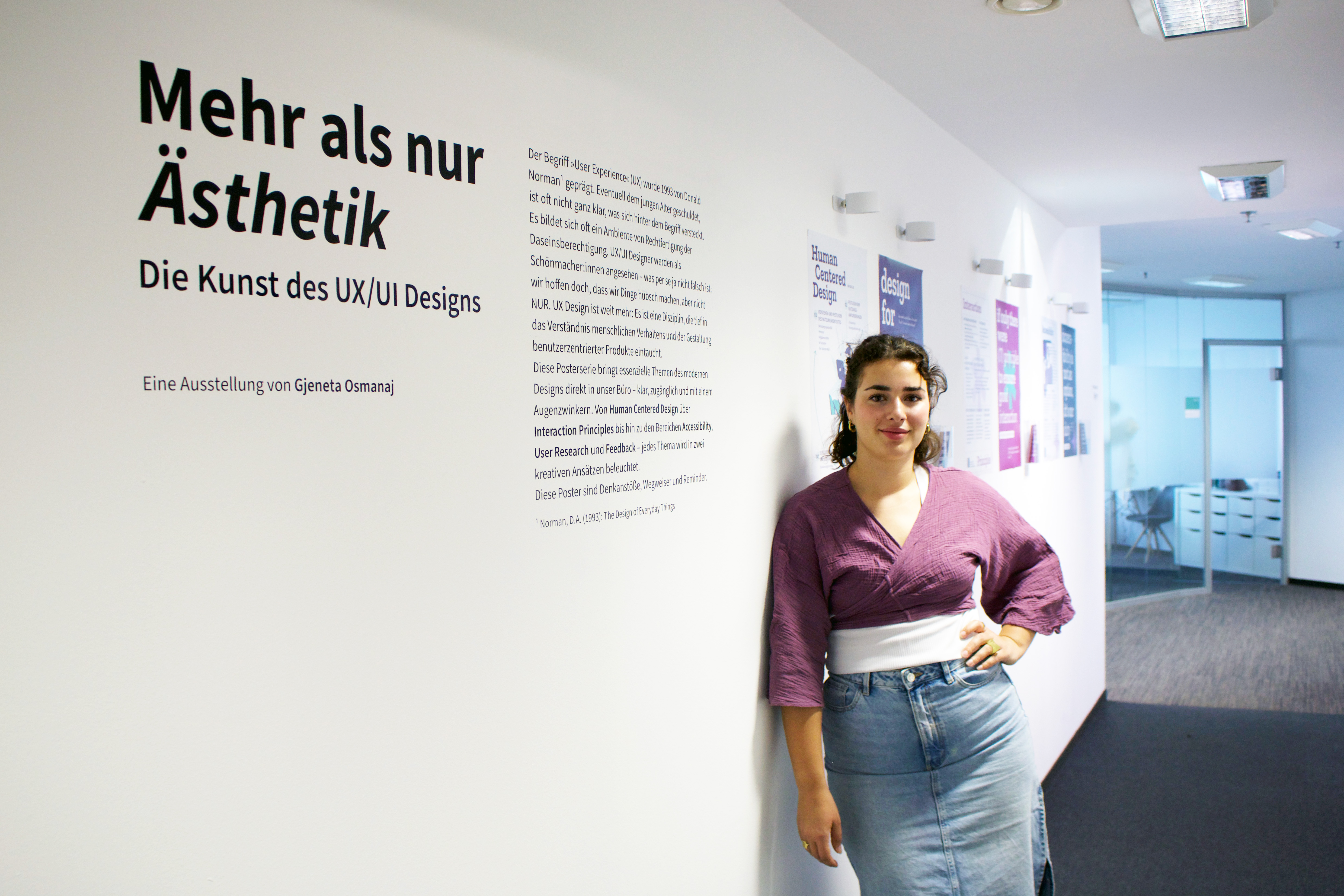 Gjeneta Osmanaj, dressed in a light denim skirt, a white T-shirt, and a purple blouse over it, is leaning against the wall of an office. She has dark curly hair, which she has tied up. Behind and in front of her, texts and posters can be seen.
