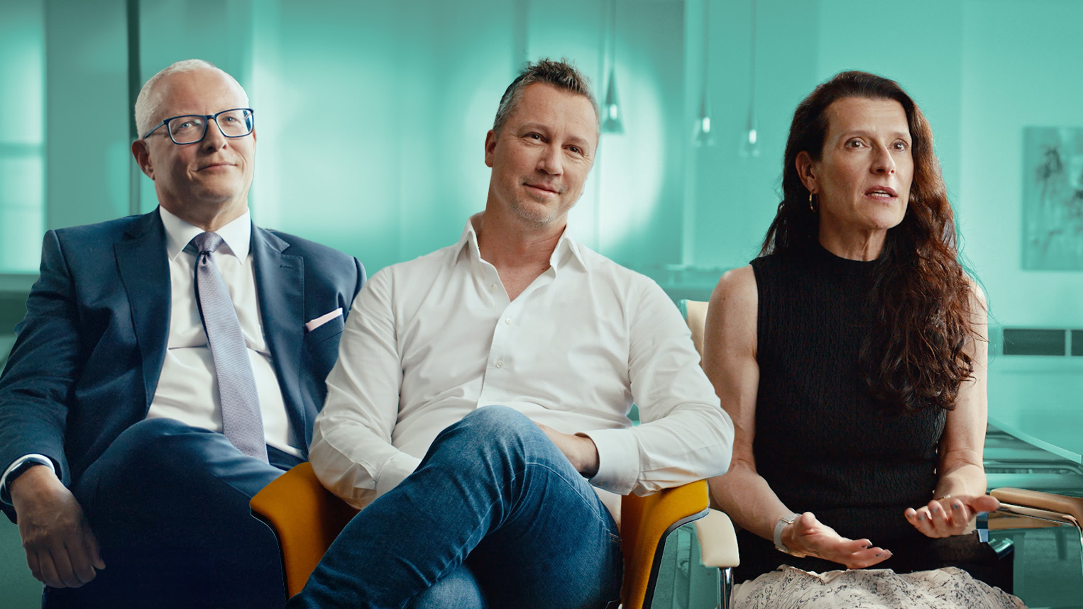 Three people — Gerhard Wagner, Thomas Kern, and Gerlinde Scholler — are seated next to each other in a modern, light-filled room. On the left, Gerhard Wagner wears a dark suit and glasses; in the middle, Thomas Kern is dressed in a white shirt and jeans; on the right, Gerlinde Scholler has long brown hair and wears a sleeveless black top. They are all looking attentively forward or to the side, with Gerlinde Scholler speaking and gesturing with her hands.