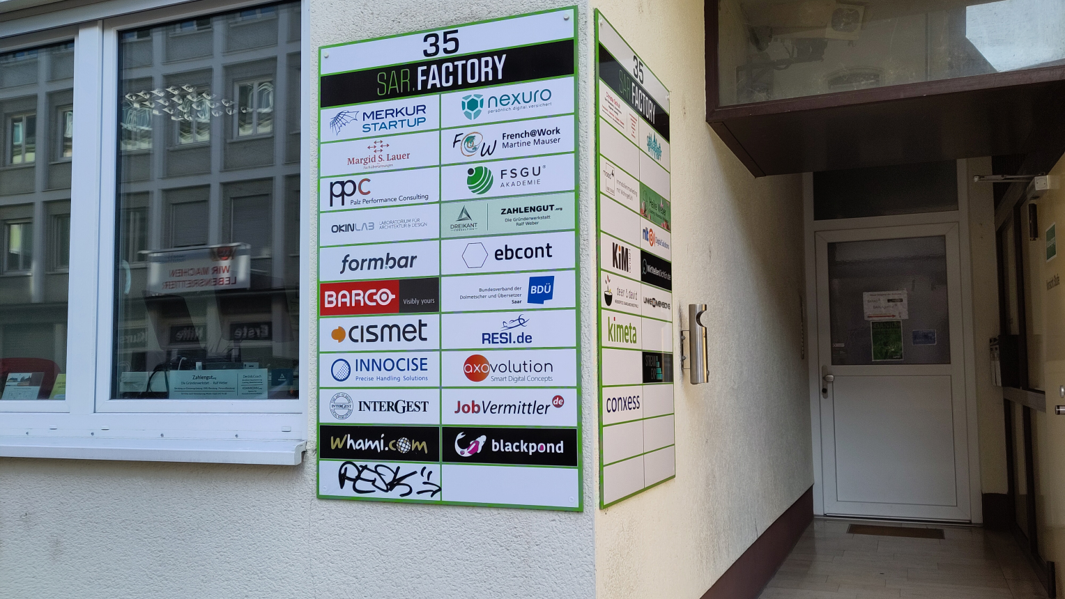 Eingangsbereich der SAR Factory in einem Geschäftsgebäude mit einem großen Schild, das die Namen und Logos mehrerer Unternehmen zeigt, darunter EBCONT, Merkur Startup, BARCO, cismet und weitere. Rechts führt ein überdachter Durchgang zu einer weißen Eingangstür mit Briefkasten und Türschild. Links spiegelt sich die gegenüberliegende Straßenseite in einem Fenster.