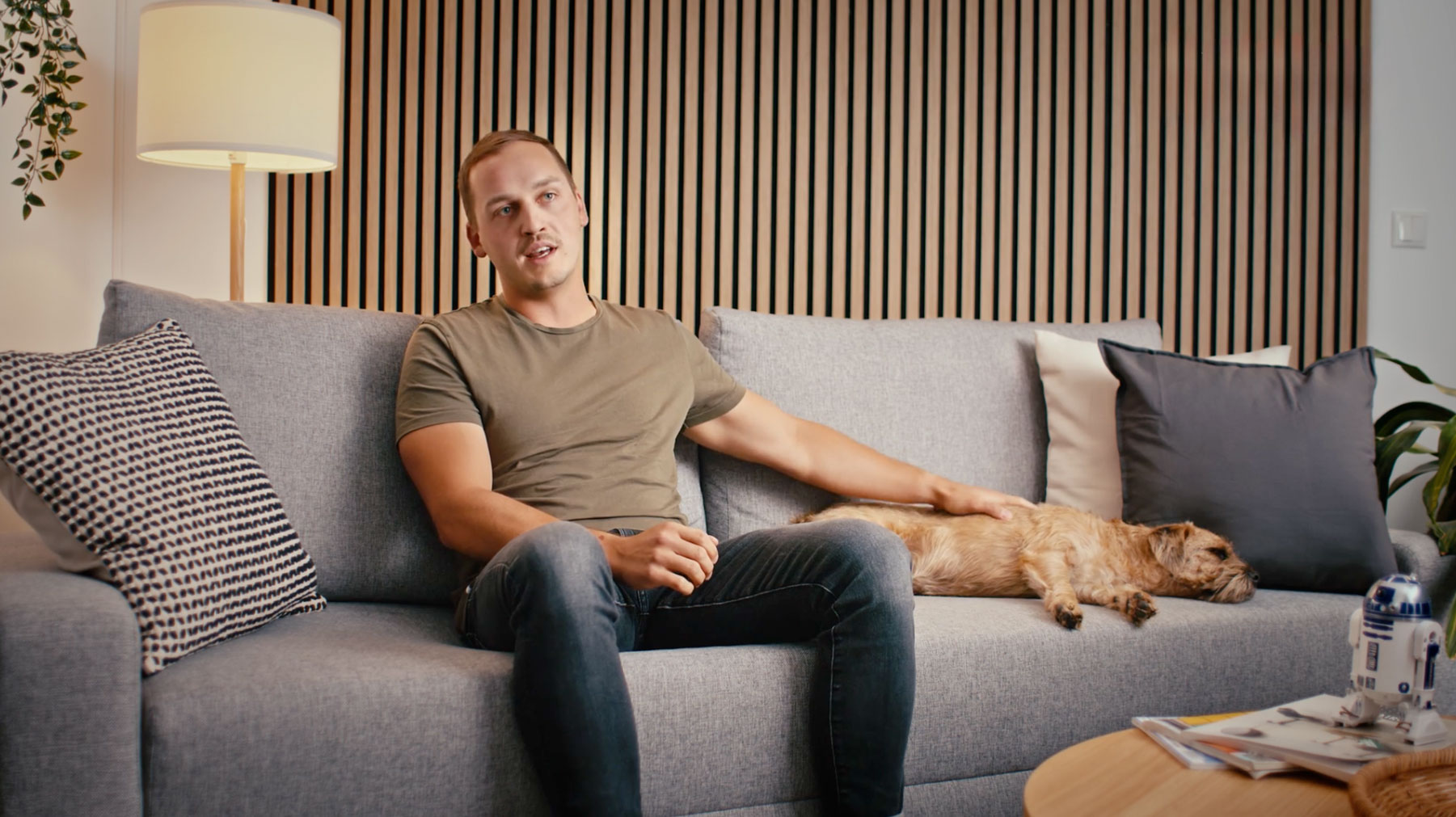 A still from the video: Patrick is sitting on a grey couch, talking and stroking his dog Otto, who is lying asleep on the couch. Patrick is wearing a brown T-shirt and dark blue jeans. The atmosphere is relaxed and it has a living room feel.