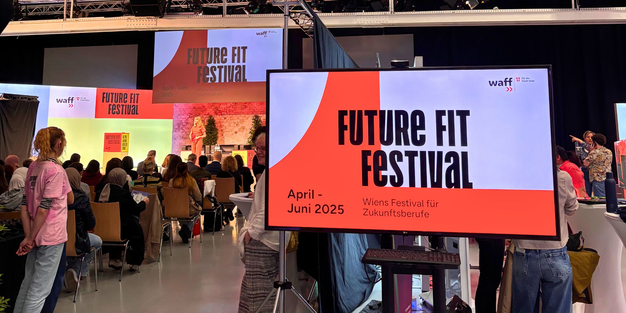 You can see a screen with the text "Future Fit Festival" and a red-purple background. A large room where women are sitting and standing. At the end, a stage with a woman speaking.