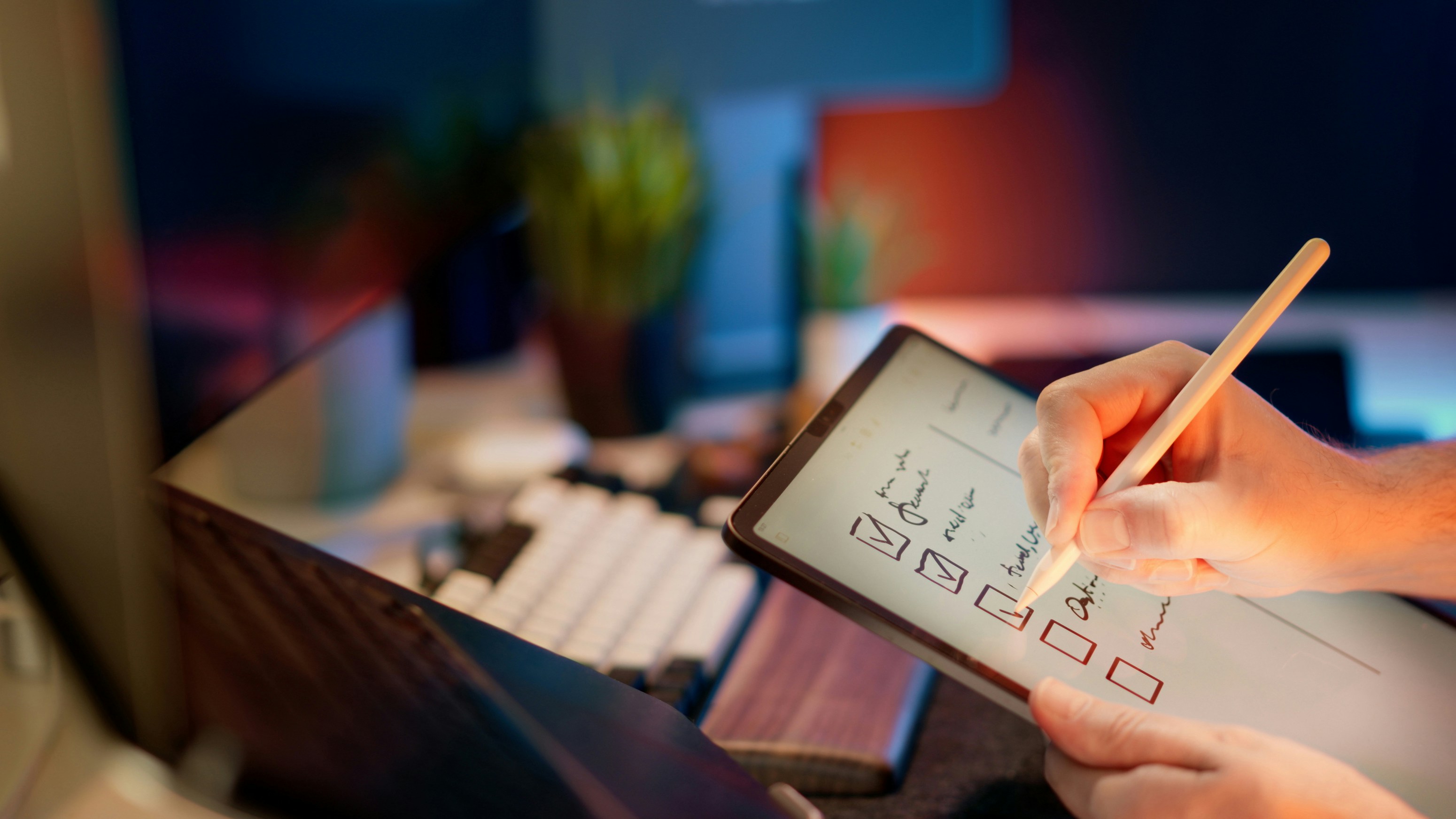 A person holds a tablet in one hand and writes on a checklist with a digital pen. Some boxes are already checked. In the blurred background, a keyboard, monitor, and plants are visible.