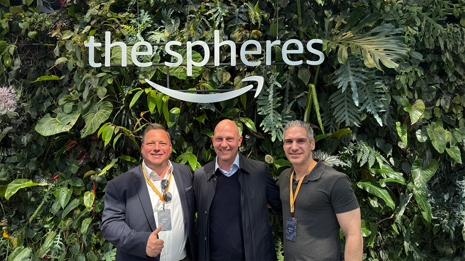 Three men stand smiling in front of a lush green plant wall with the large white sign “the spheres” and the Amazon arrow logo. Two of them wear guest badges on lanyards around their necks.