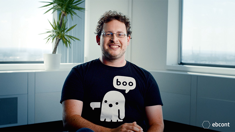 A smiling man with glasses and curly hair sits in front of a window in a modern office. He is wearing a black T-shirt with a white cartoon ghost and the speech bubble “boo”. The EBCONT logo is visible at the bottom right of the picture.
