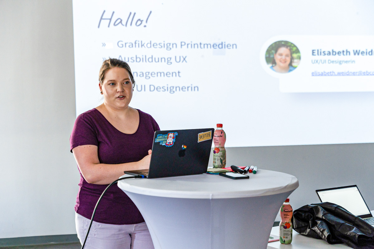 Elisabeth Weidner is standing at a high table, giving a presentation. Behind her, a slide is projected displaying information about her background as a UX/UI designer. On the table are a laptop with stickers, bottles of iced tea, markers, and a smartphone.