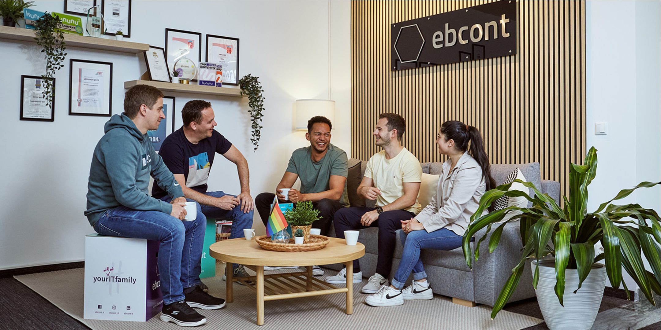 Mehrere Personen sitzen zusammen auf einem Sofa und plaudern. Dahinter eine braune Holzwand und das EBCONT Logo