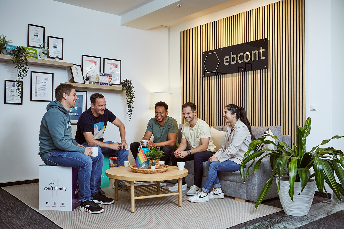 Five people sit laughing and chatting in a cozy lounge corner at EBCONT. There is a small rainbow flag on the table. The atmosphere is open, friendly and appreciative.