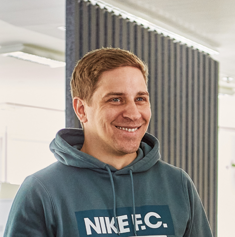 A man with light brown hair stands in half profile in front of a partition wall in an office