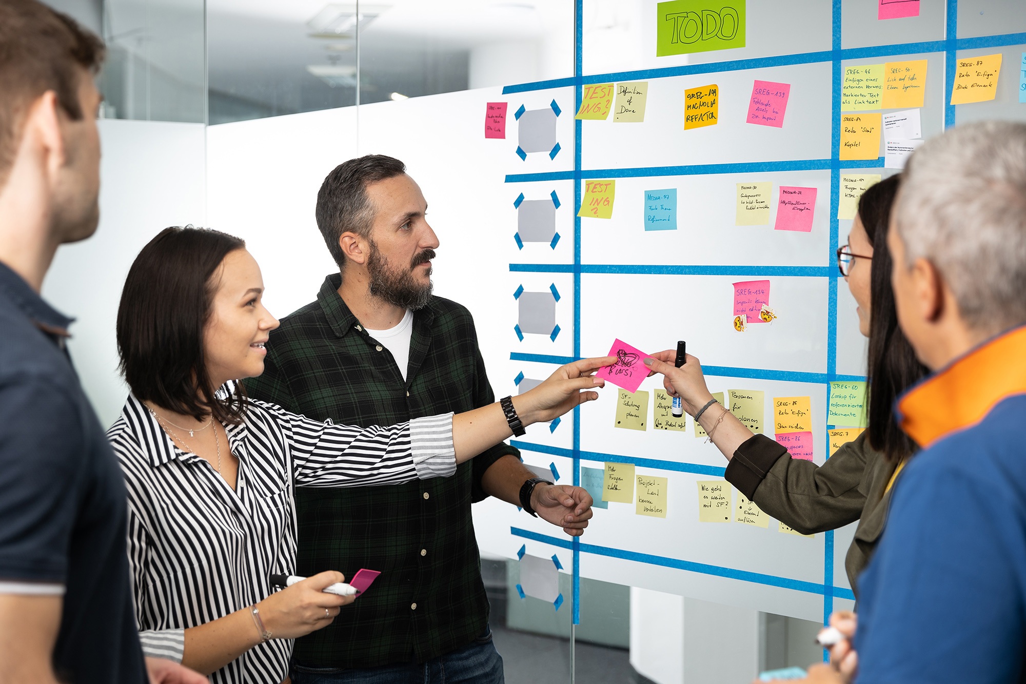 A team discusses tasks in front of a large Kanban board with colored sticky notes. Several people are pointing to individual cards and actively discussing. The scene shows collaborative work in an agile project environment.