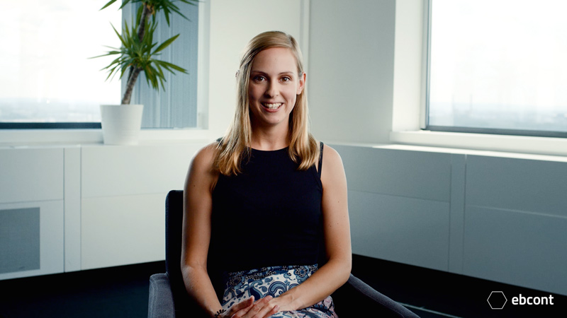 A frame from the video showing employee Katharina sitting relaxed in a bright room and laughing at the camera. She is wearing a black top.